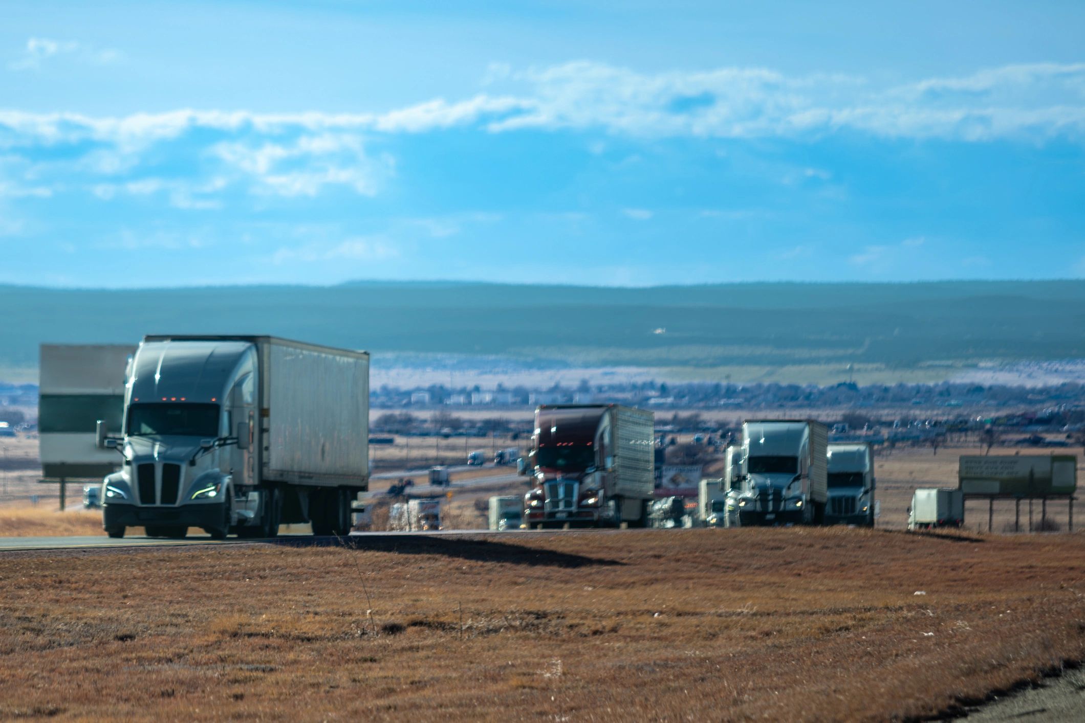 Lineup of commercial trucks parked on arid plateau overlooking distant city. Multiple box and tanker vehicles staged in rural Colorado high desert landscape.
